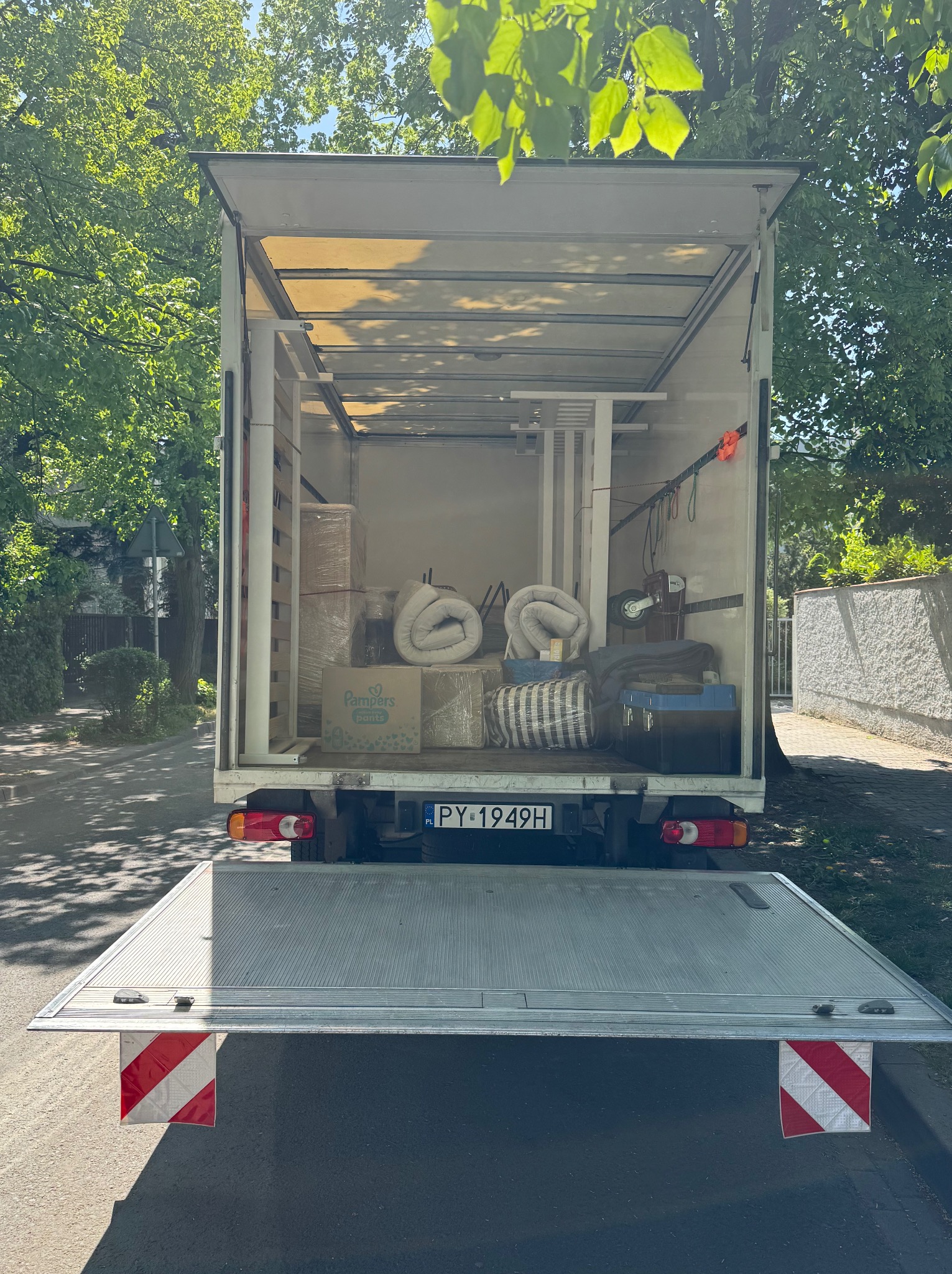 Otwarta paka busa załadowana kartonami, meblami i zwiniętymi materacami, gotowa do transportu. Widoczny numer rejestracyjny pojazdu na tle zieleni.