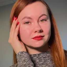 Portret kobiety o rudych włosach z czerwonymi ustami i paznokciami, dłoń przy twarzy. Subtelny makijaż oczu i brwi. Szary sweter i jasne tło.