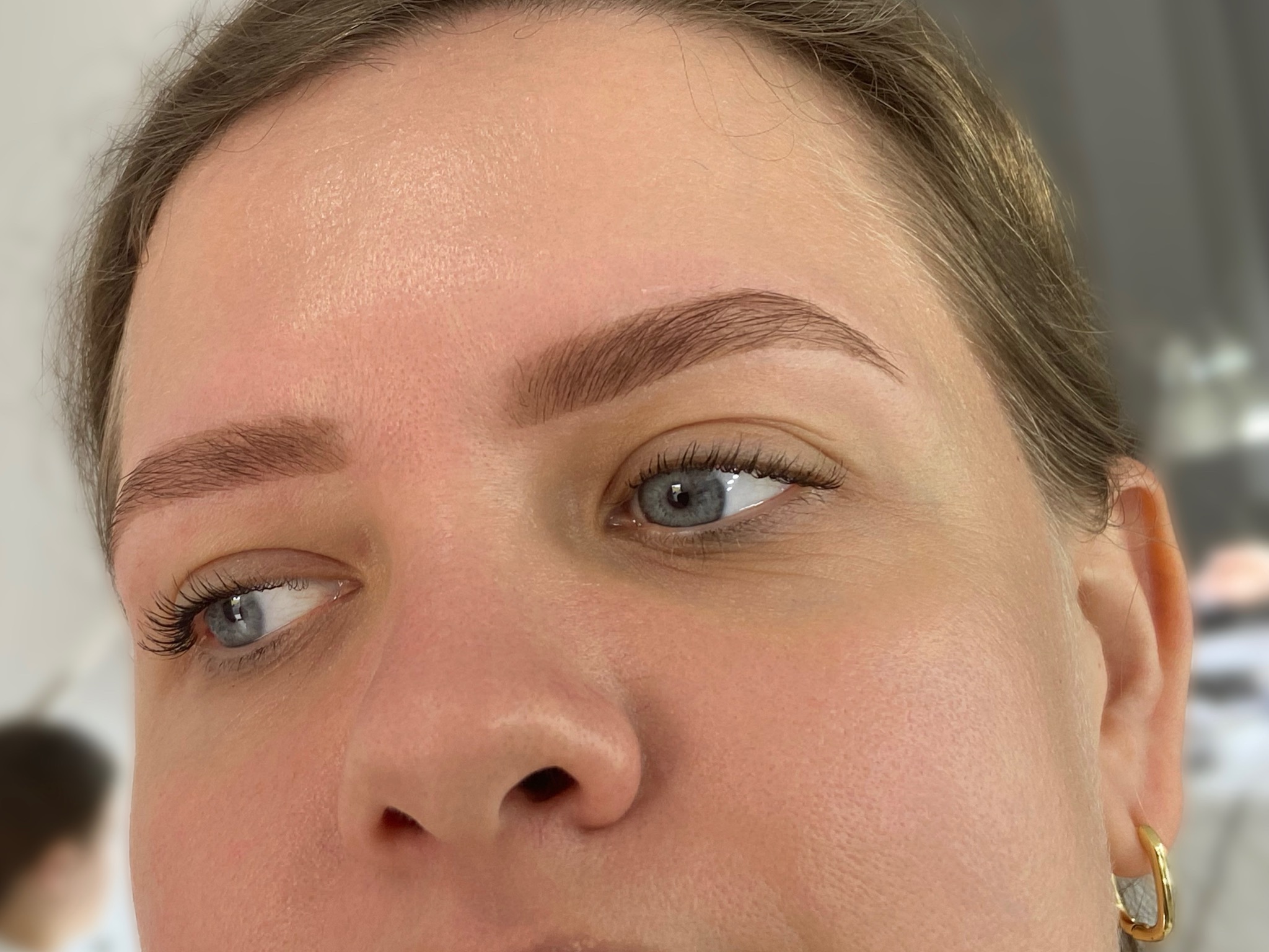 Zbliżenie na twarz kobiety z delikatnym makijażem brwi i rzęs. Widoczne złote kolczyki i niebieskie oczy. Naturalne oświetlenie.