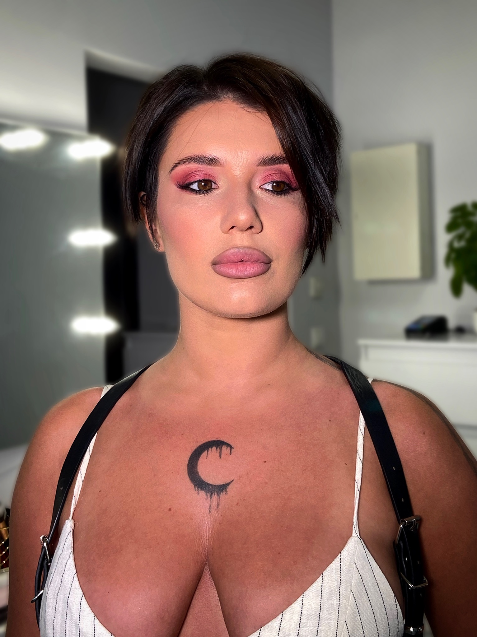 Portret kobiety z profesjonalnym makijażem oczu w odcieniach różu i podkreślonymi ustami. Widoczny tatuaż na dekolcie w kształcie księżyca.