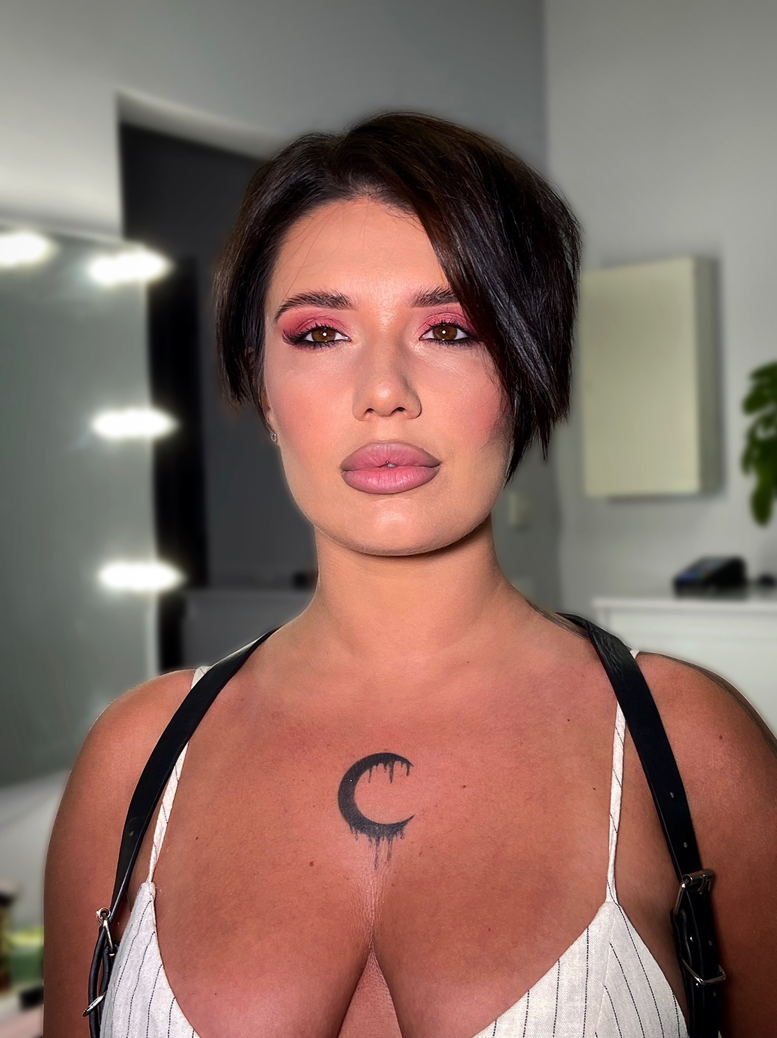 Portret kobiety z profesjonalnym makijażem oczu w odcieniach różu i podkreślonymi ustami w studiu wizażu, z tatuażem półksiężyca na dekolcie.