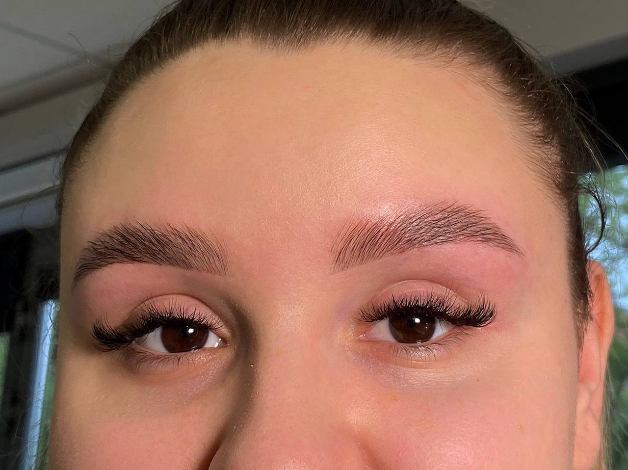 Zbliżenie na twarz kobiety po zabiegu laminacji brwi i przedłużania rzęs. Widoczne brązowe oczy i idealnie ułożone, ciemne brwi.