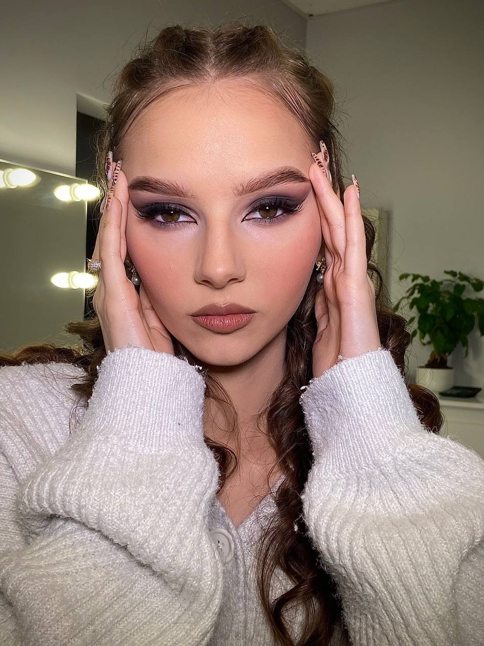 Portret kobiety z profesjonalnym, wieczorowym makijażem oczu w odcieniach brązu i fioletu, dłonie przy twarzy, z widocznymi zdobieniami na paznokciach.