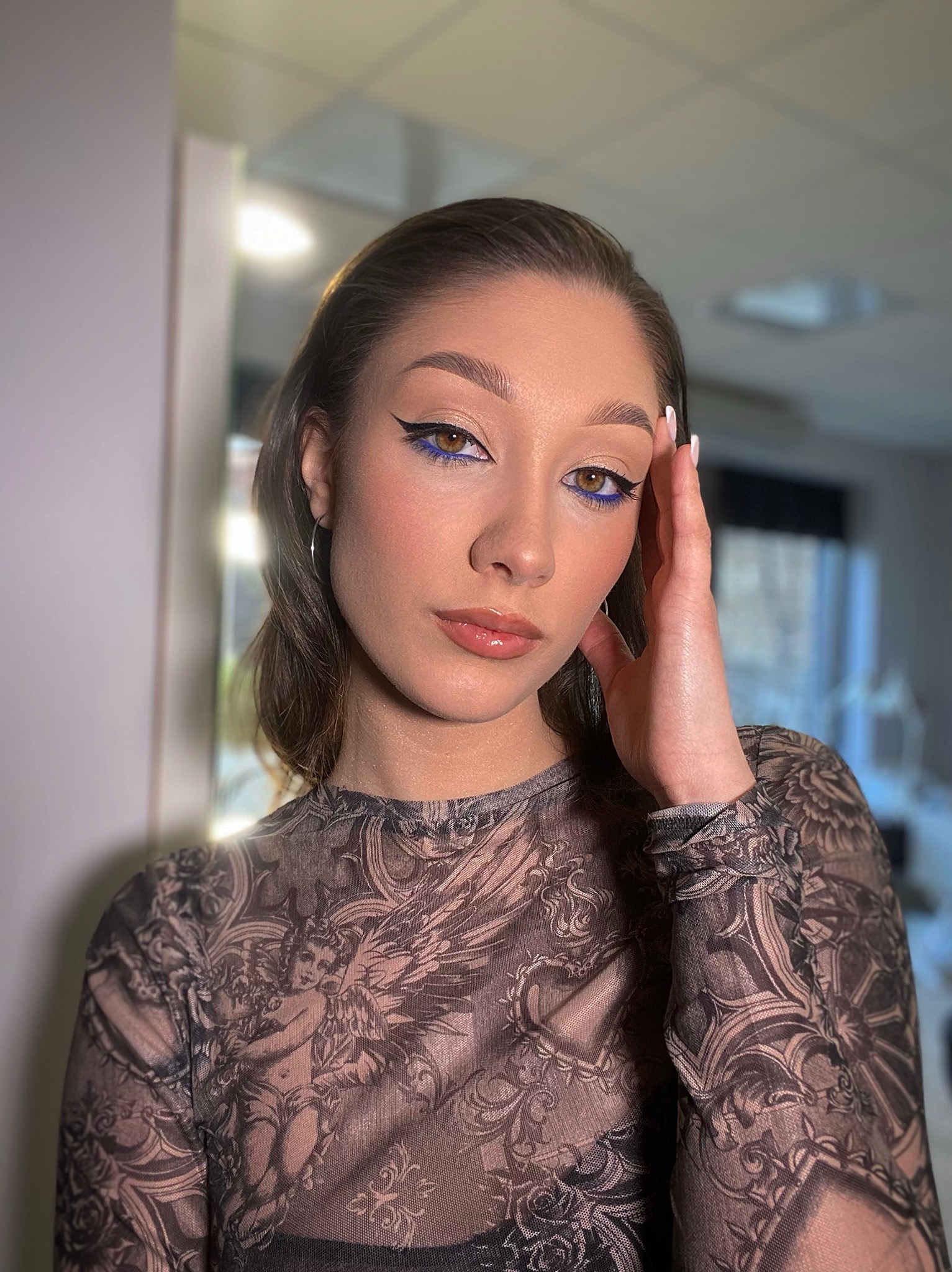 Portret kobiety z wyrazistym, niebieskim eyelinerem i błyszczącymi ustami. Uwagę przyciąga wzorzysta, prześwitująca bluzka i delikatna biżuteria.