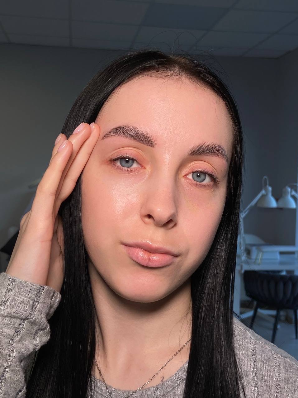 Portret kobiety z idealnie wystylizowanymi brwiami, podkreślonymi niebieskimi oczami i delikatnym makijażem ust. Długie, czarne włosy opadają na ramiona.