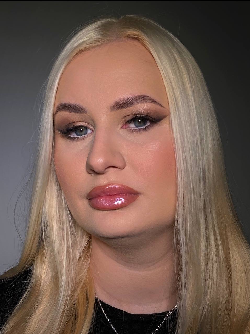 Portret blondynki z precyzyjnym makijażem oczu (eyeliner, cienie) i błyszczącymi ustami, na neutralnym tle.