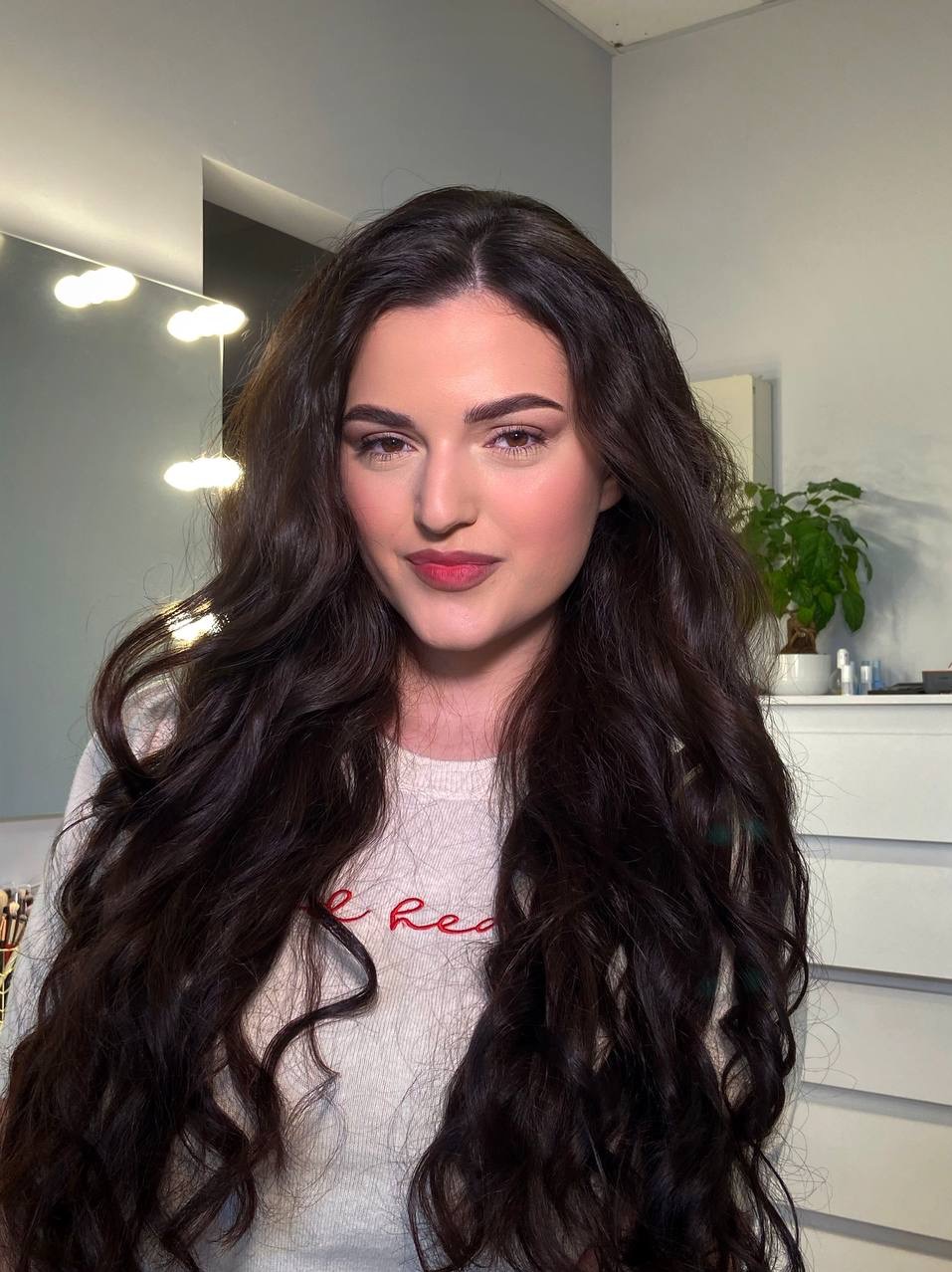 Portret brunetki z delikatnym makijażem w odcieniach różu, prezentujący efekt końcowy usługi w salonie. W tle toaletka z oświetleniem i roślina doniczkowa.