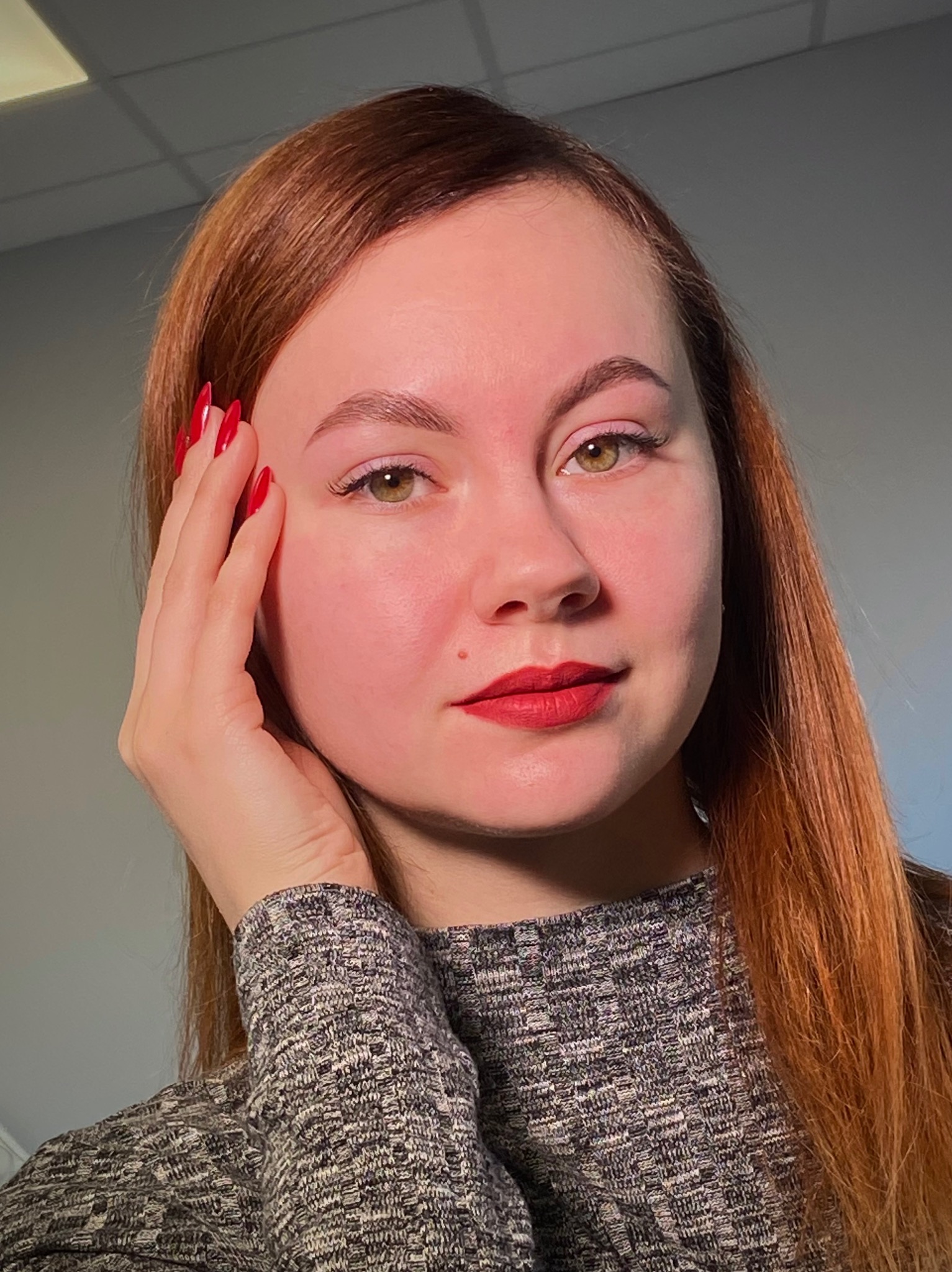 Portret kobiety o rudych włosach z czerwonymi ustami i paznokciami, dłoń przy twarzy. Subtelny makijaż oczu i brwi. Szary sweter i jasne tło.