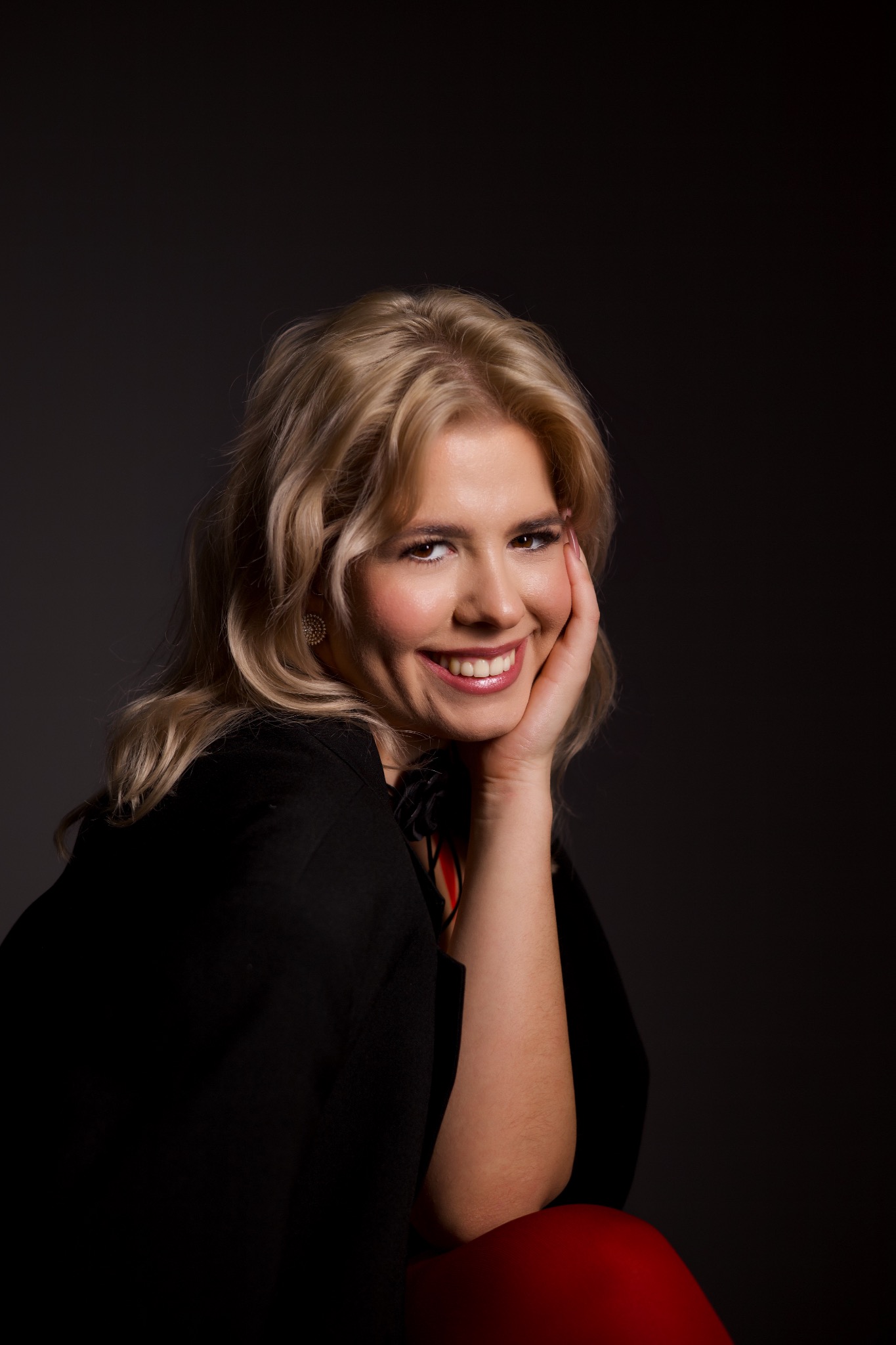 Uśmiechnięta blondynka w czarnej marynarce, z delikatnym makijażem i błyszczącymi kolczykami, pozuje na ciemnym tle, opierając twarz na dłoni.