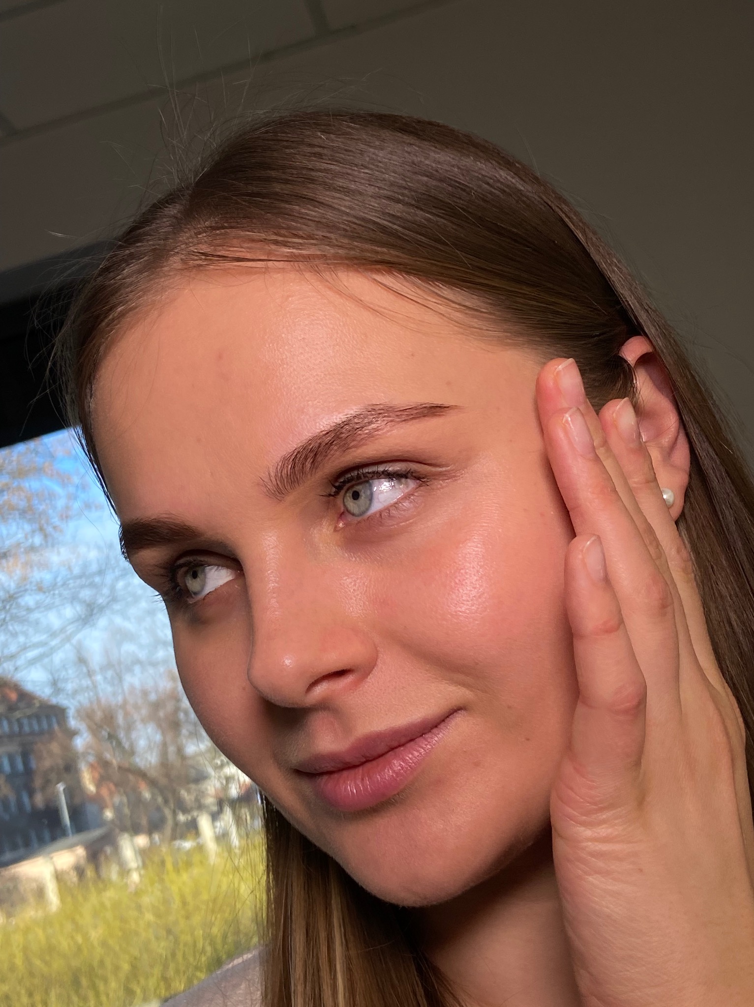 Portret kobiety z delikatnym makijażem, podkreślone brwi i naturalny blask cery. Widok z bliska, dłoń przy twarzy, w tle okno z widokiem na budynek.