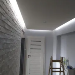 Wykończone wnętrze z oświetleniem LED przy suficie, ściana z białej cegły, drewniane drzwi i drabina malarska w rogu. Widok na korytarz po remoncie.