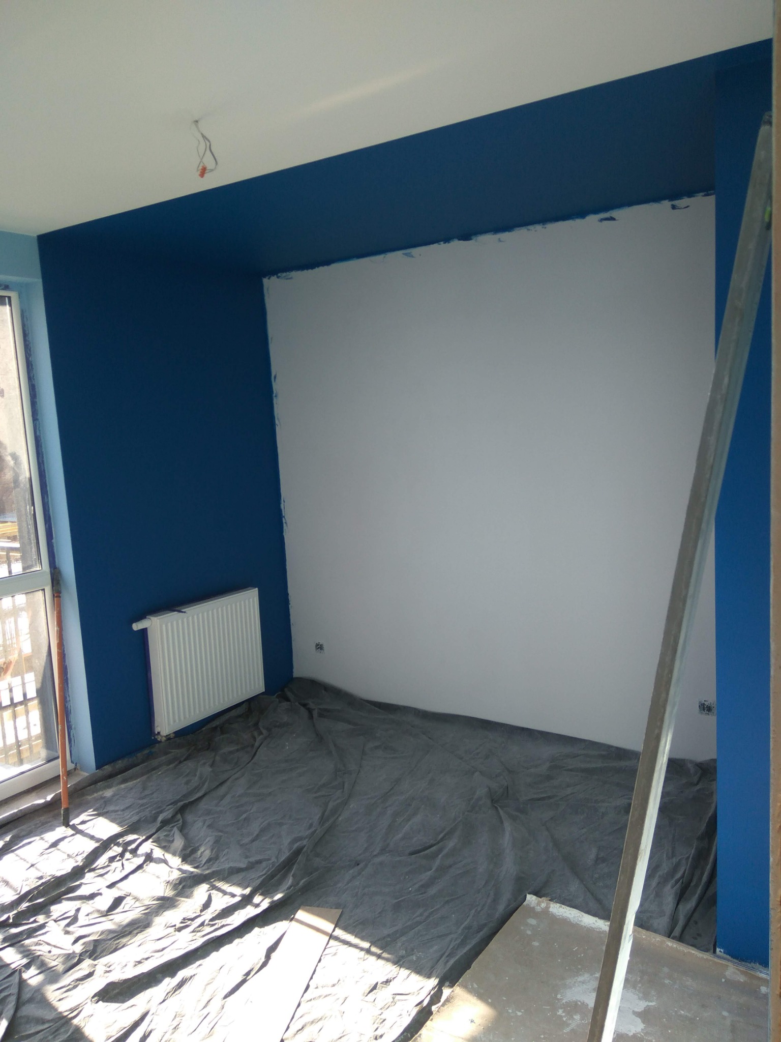 Pokój w trakcie malowania: ściany w błękicie i bieli, podłoga zabezpieczona folią, widoczny grzejnik i fragment drabiny malarskiej.