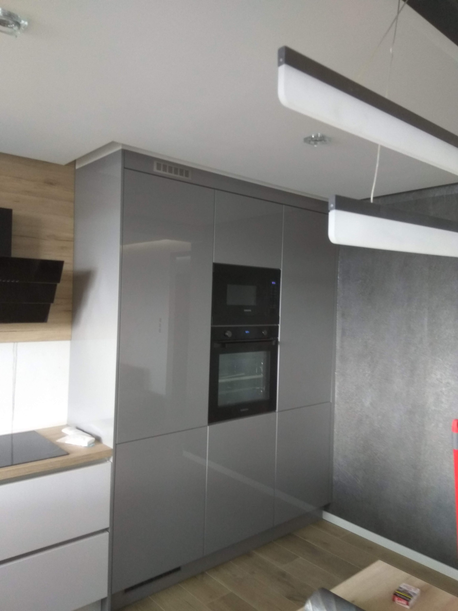 Nowoczesna kuchnia w szarościach: zabudowa meblowa z piekarnikiem i mikrofalówką, minimalistyczny design, drewniane akcenty blatu i podłogi, oświetlenie ledowe.