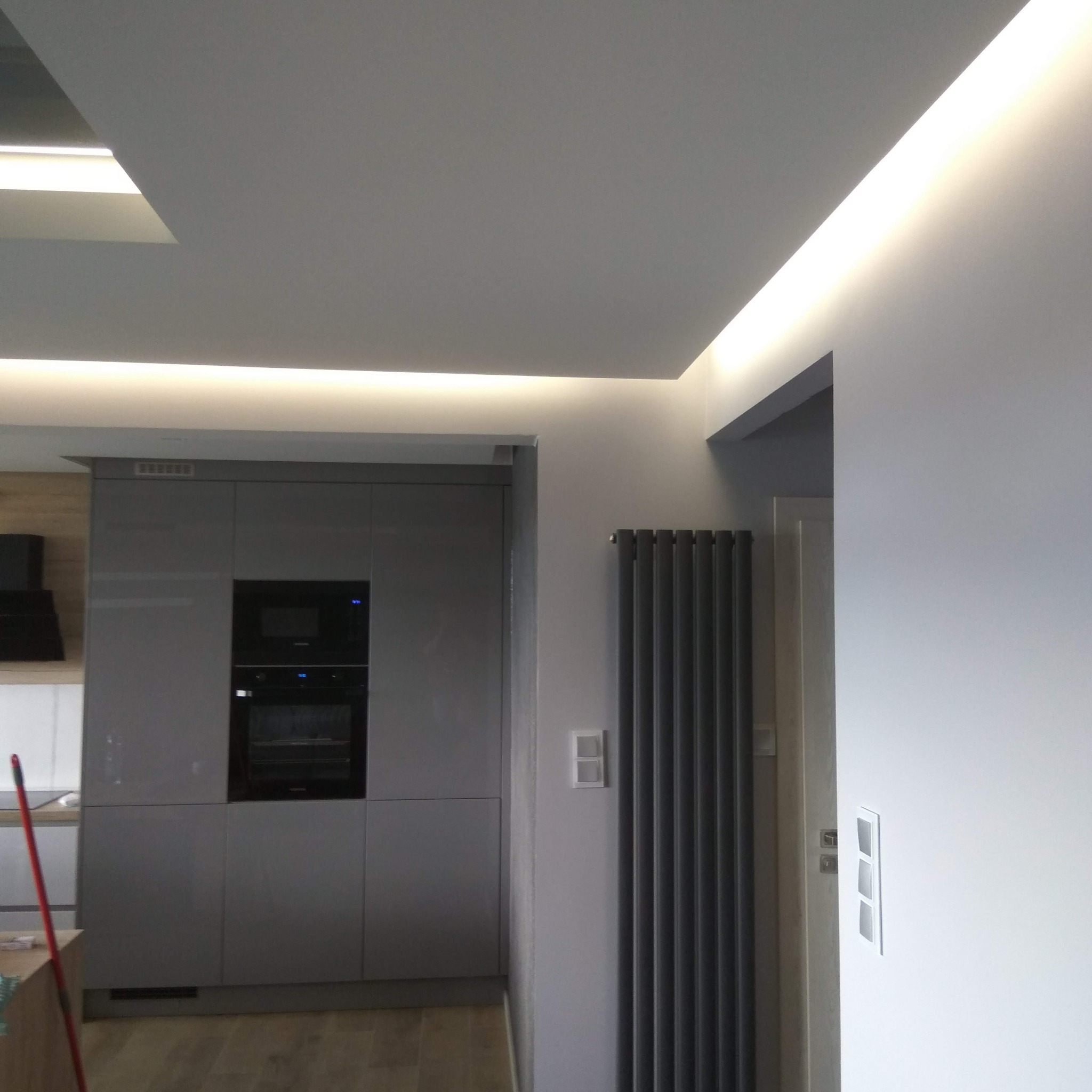 Szare, nowoczesne wnętrze: zabudowa kuchenna z piekarnikiem, grzejnik panelowy i podświetlany sufit. Minimalistyczny design.