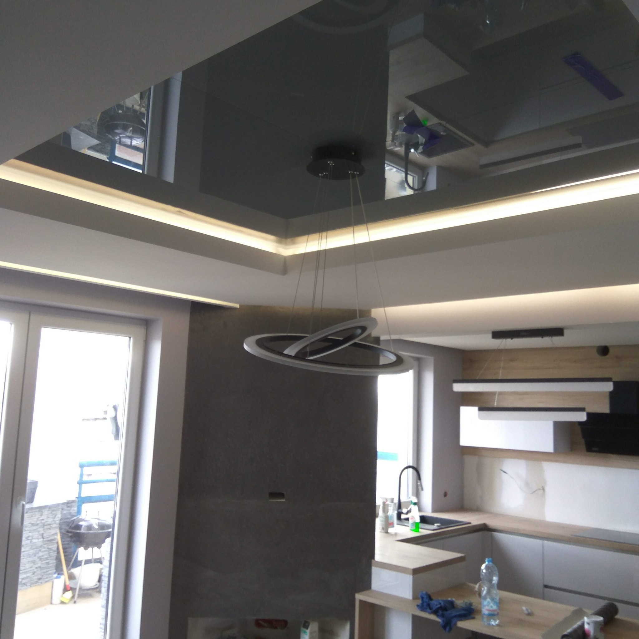 Nowoczesne wykończenie wnętrza: błyszczący sufit z oświetleniem LED, designerska lampa wisząca, fragment kuchni z jasnymi szafkami i szara ściana.