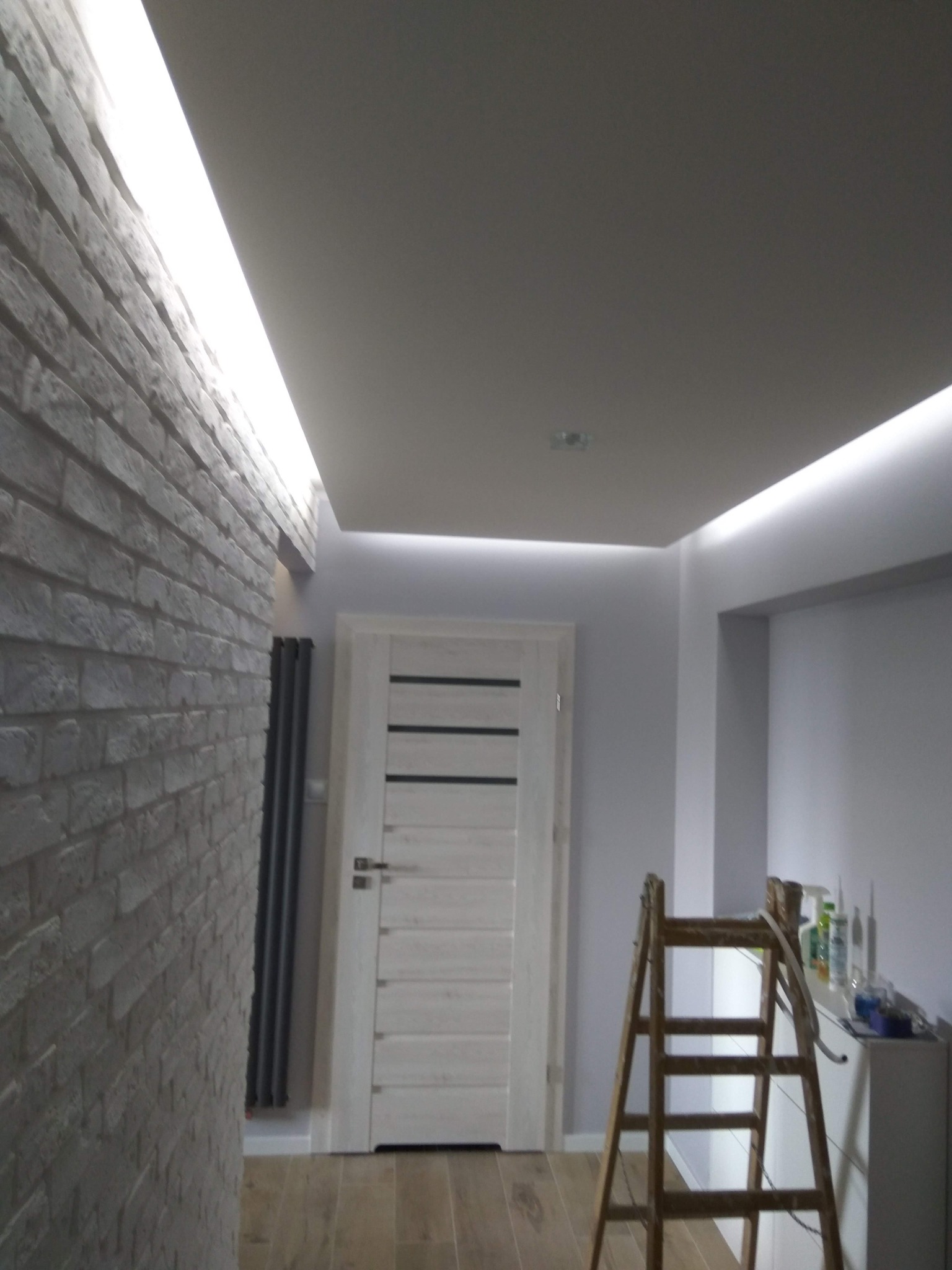 Wykończone wnętrze z oświetleniem LED przy suficie, ściana z białej cegły, drewniane drzwi i drabina malarska w rogu. Widok na korytarz po remoncie.
