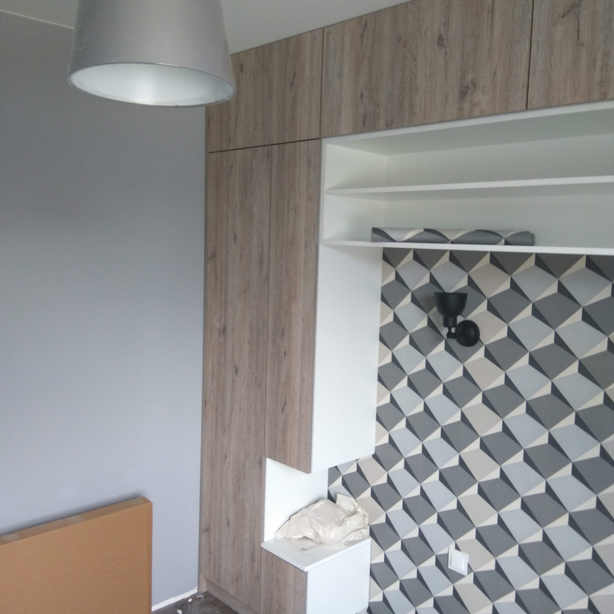 Wykończona garderoba z drewnopodobnymi frontami, białymi półkami i geometryczną tapetą w szarościach. Widoczna lampa sufitowa i fragment podłogi.