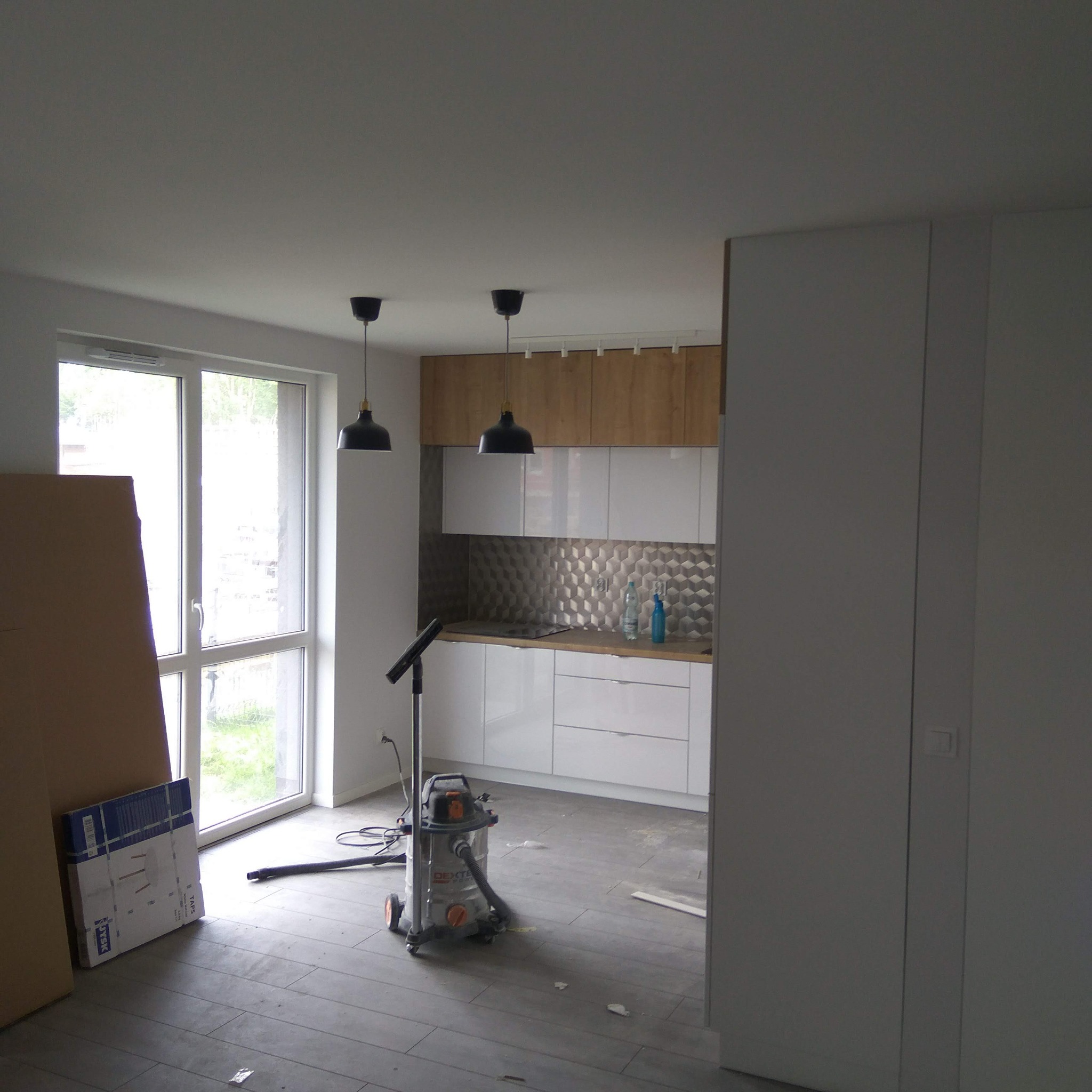 Wnętrze w trakcie wykańczania: widoczna kuchnia z szarą mozaiką, podłoga z płytek imitujących drewno, odkurzacz przemysłowy i okno.