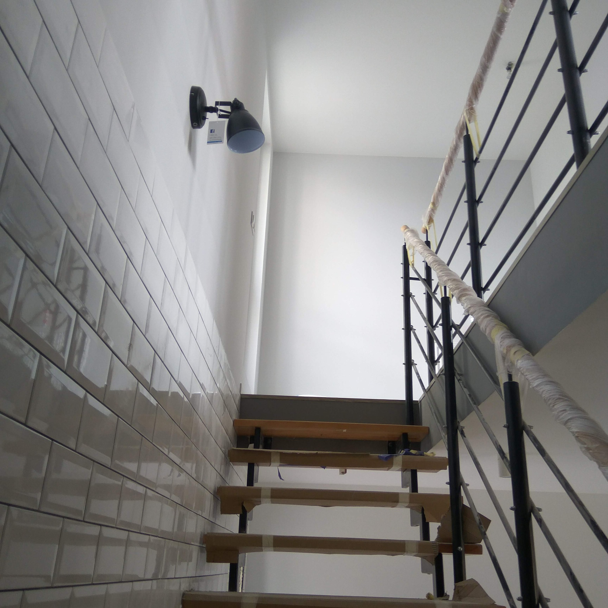 Schody z drewnianymi stopniami w trakcie wykańczania, zabezpieczone taśmą. Na ścianie białe, błyszczące płytki i czarna lampa. Metalowa barierka zabezpieczona folią.