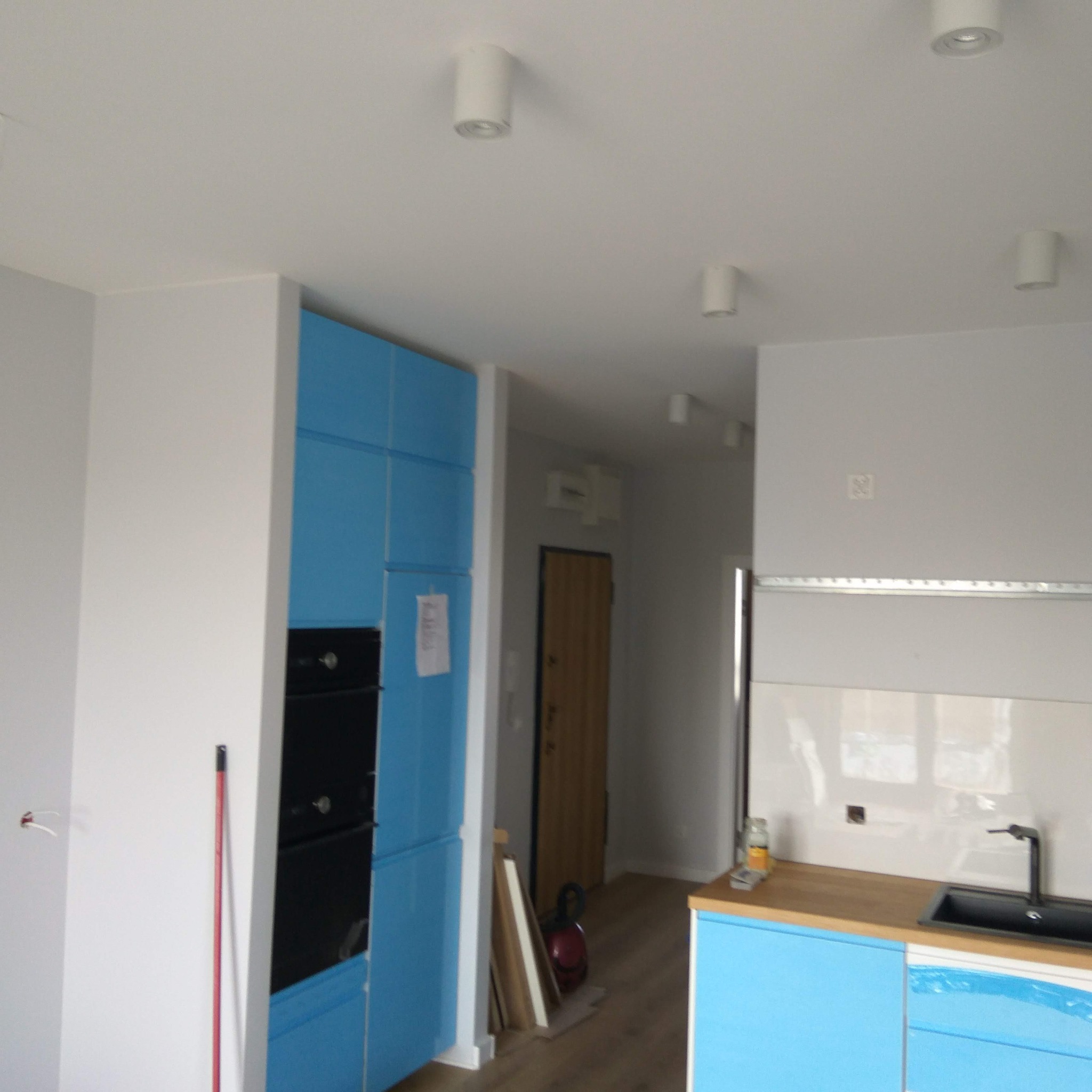 Jasne wnętrze z błękitnymi szafkami kuchennymi i białymi ścianami. Widoczne oświetlenie sufitowe i fragment drzwi wejściowych. Nowoczesny design.