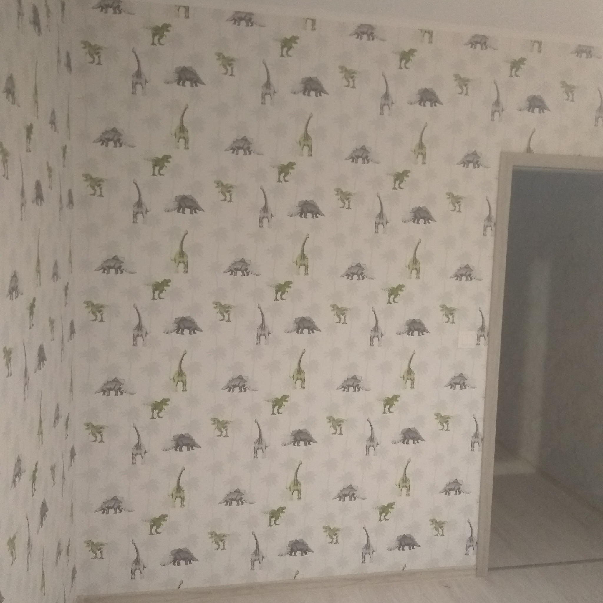 Ściana w pokoju dziecięcym z tapetą w dinozaury, widoczny fragment drzwi. Pastelowe kolory, stonowane oświetlenie.