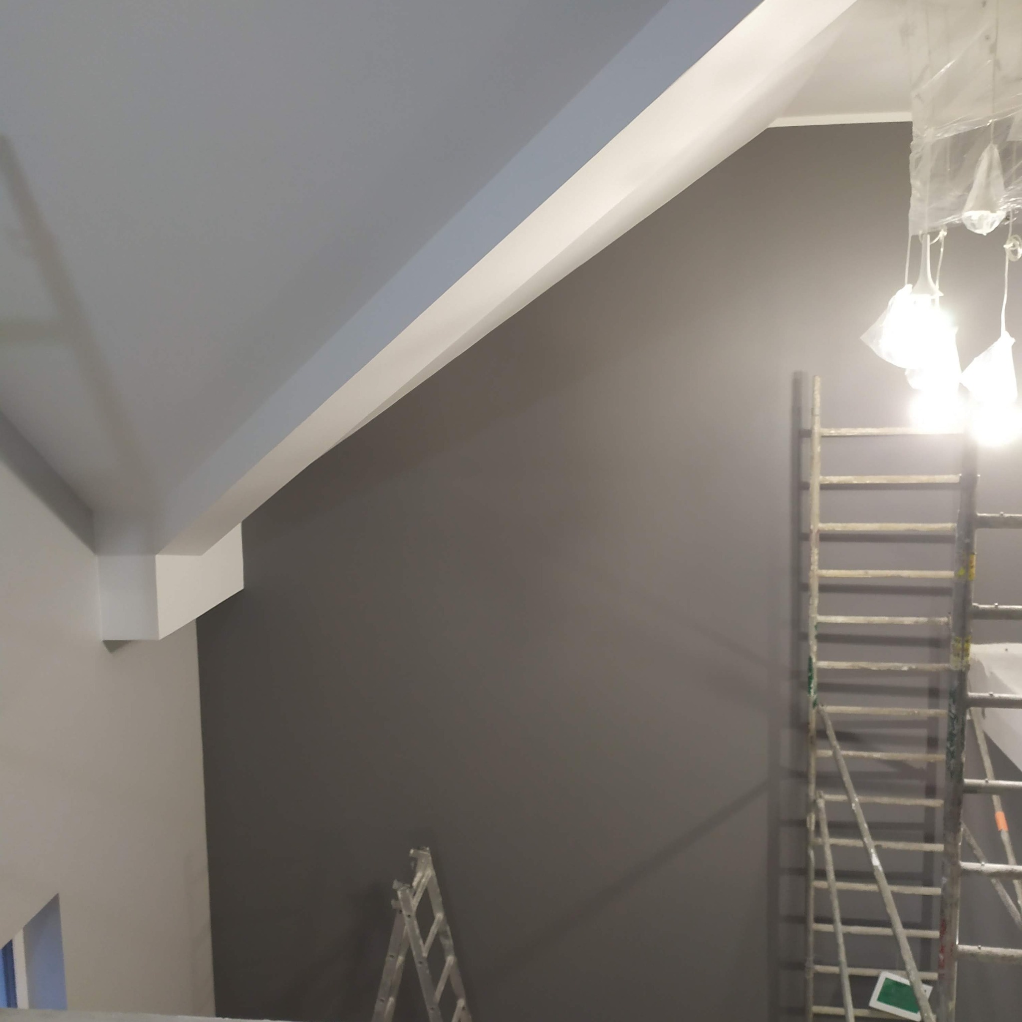 Ściana po malowaniu na szaro w pokoju ze skosami. Widoczna drabina malarska i zabezpieczone oświetlenie. Jasne listwy sufitowe.