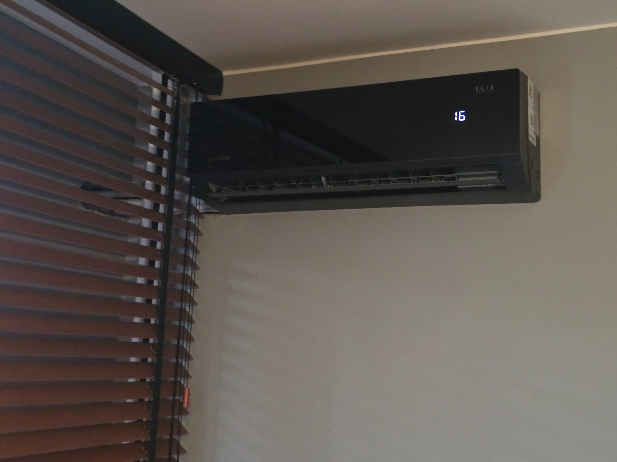 Czarna klimatyzacja ELIS zamontowana na szarej ścianie obok drewnianych żaluzji. Na wyświetlaczu widoczna temperatura 16 stopni.