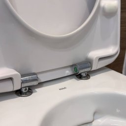 Przytulny dom - Biała, ceramiczna toaleta marki Koło z podniesioną deską, widok z bliska na zawiasy i wnętrze muszli. Czysta powierzchnia, gotowa do użytku.