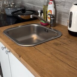 Przytulny dom - Błyszczący, czysty zlew w kuchni z drewnianym blatem, obok którego stoją płyn do naczyń, butelka z olejem i czajnik. Widoczne garnki na płycie.
