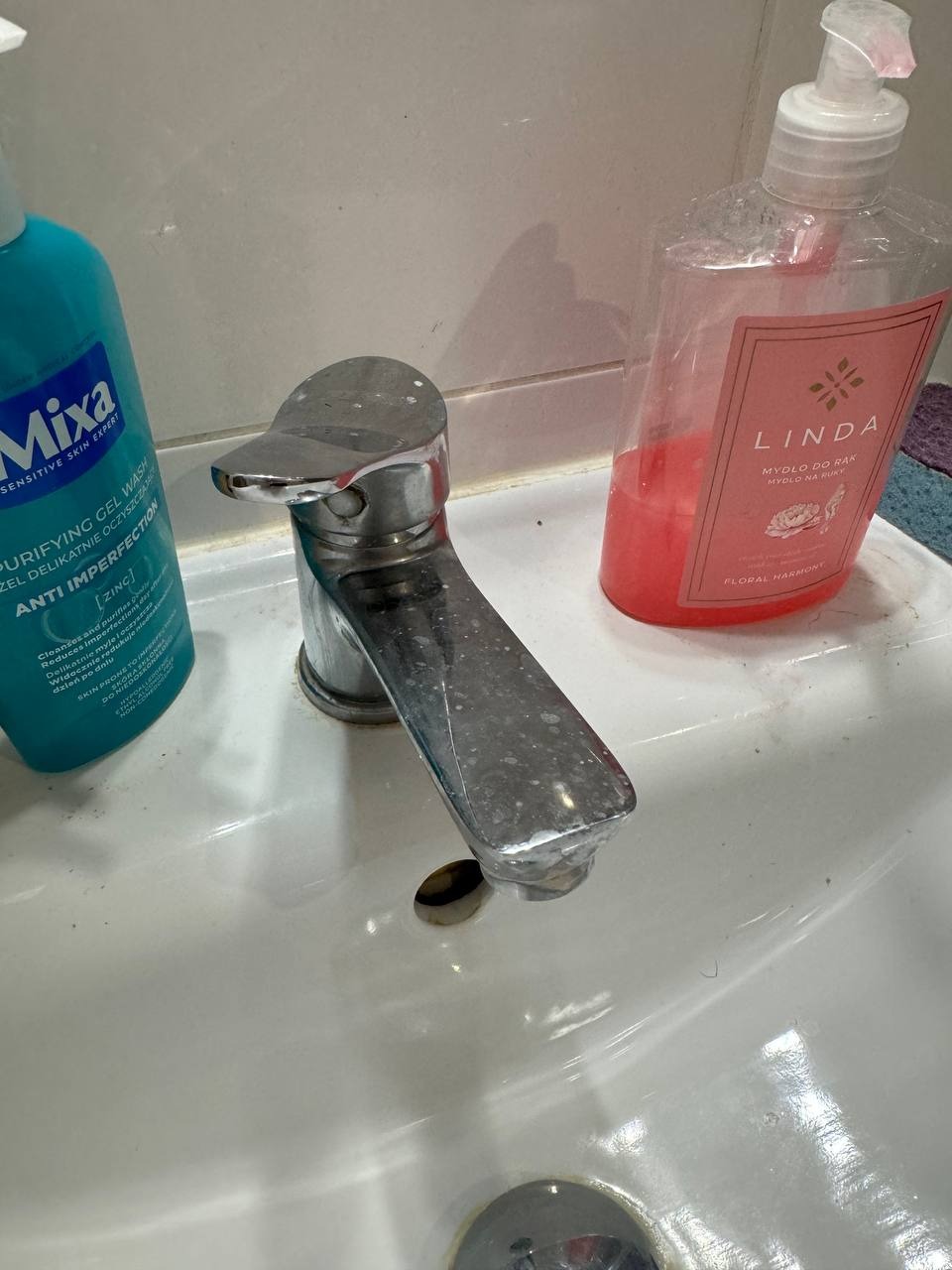 Zlew z mydłem w płynie i żelem do mycia twarzy. Widoczne ślady użytkowania na armaturze i zlewie. Ujęcie z bliska, skupione na baterii i otoczeniu.