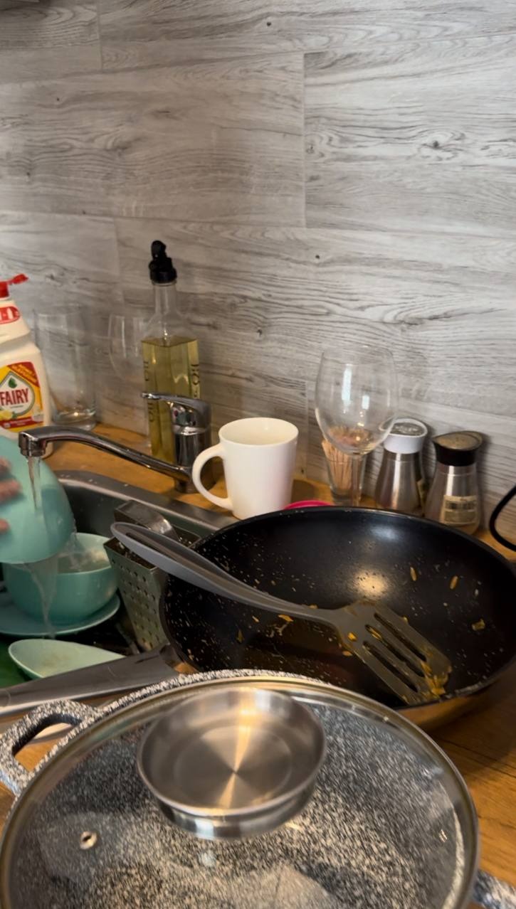 Zlew pełen naczyń, woda leje się z kranu na talerz. Widoczne garnki, patelnie, szklanki i płyn do naczyń. Zabrudzenia na naczyniach.