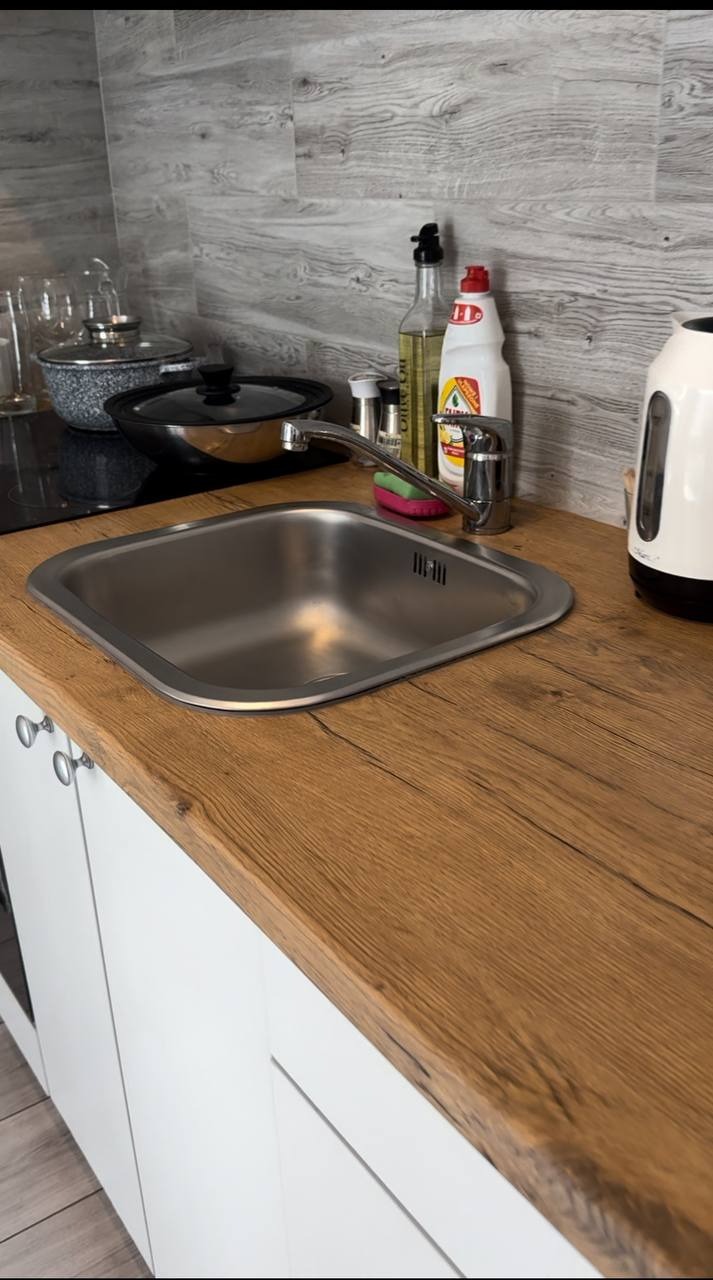 Błyszczący, czysty zlew w kuchni z drewnianym blatem, obok którego stoją płyn do naczyń, butelka z olejem i czajnik. Widoczne garnki na płycie.