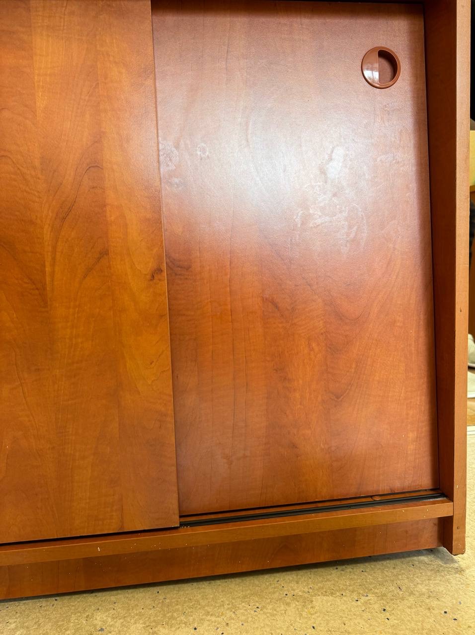 Czysta, drewniana szafka z przesuwnymi drzwiami, widoczny okrągły uchwyt. Ujęcie z dołu, ukazujące dolną krawędź i fakturę podłoża.