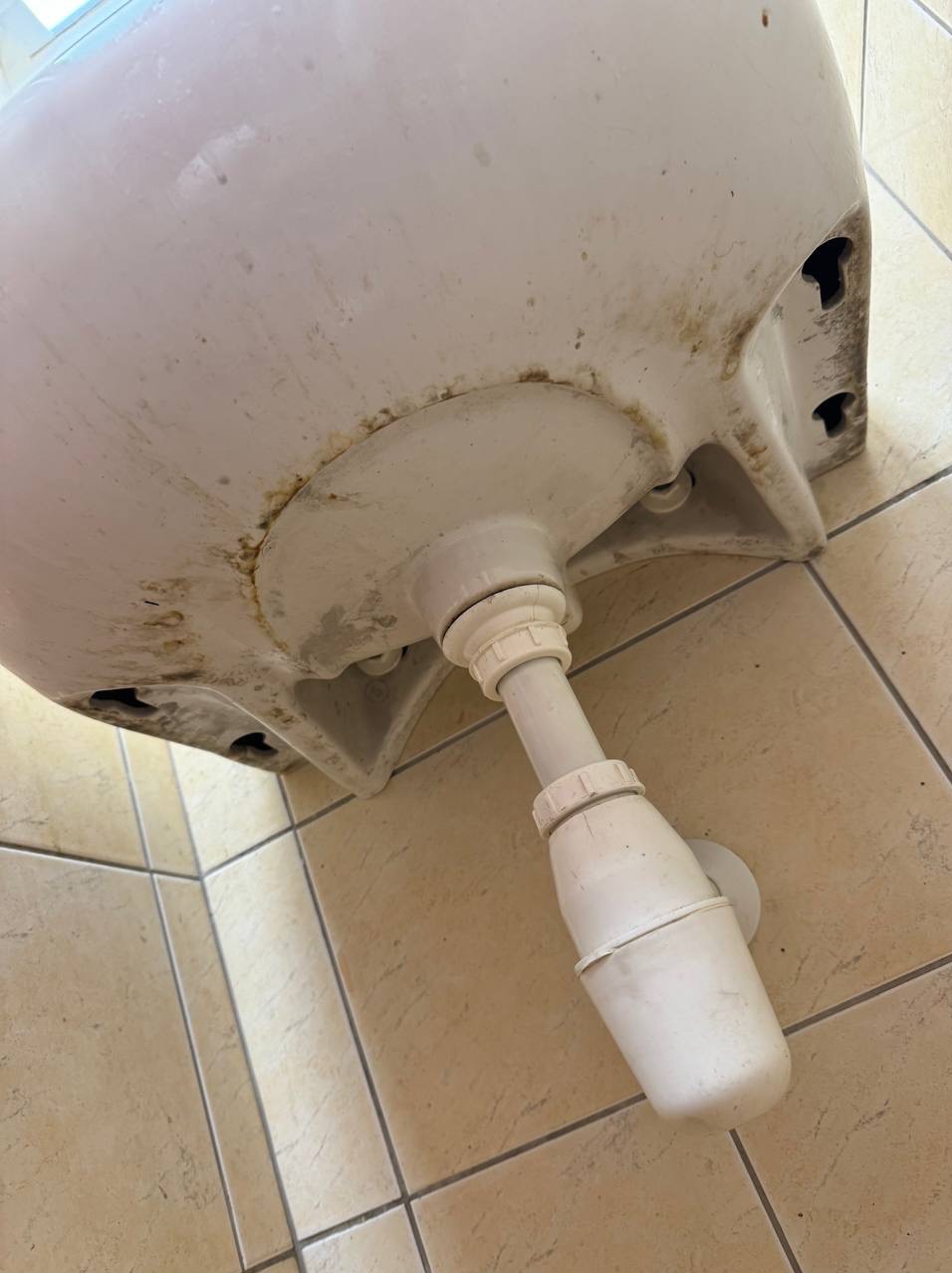 Z bliska: zabrudzona umywalka z widocznym syfonem i osadami, ukazana pod nietypowym kątem na tle płytek podłogowych. Detal higieny zaniedbanej łazienki.