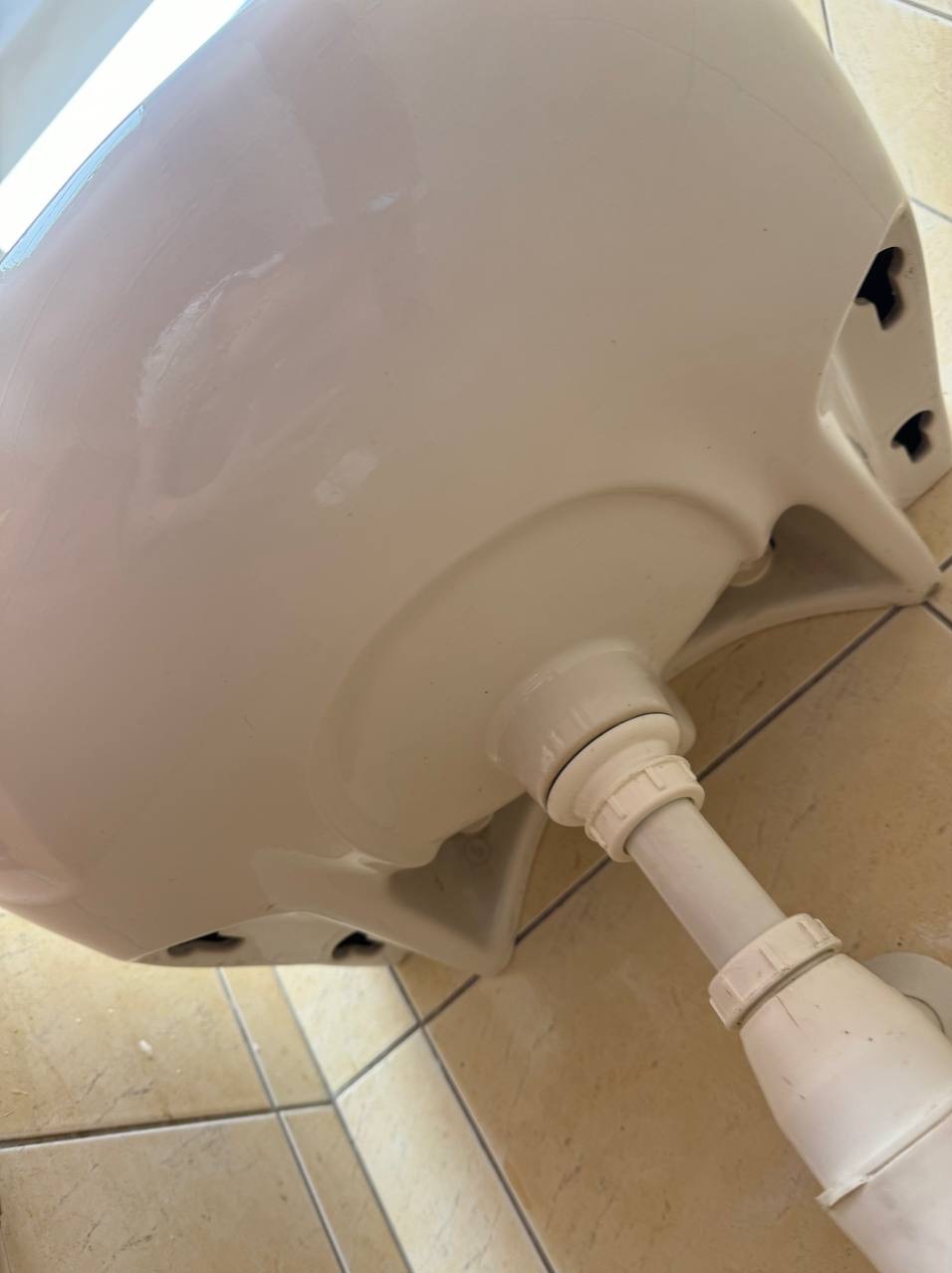 Spód umywalki z podłączonym syfonem, widok z dołu na płytki podłogowe. Biała ceramika i plastikowe rury. Zdjęcie z bliska.