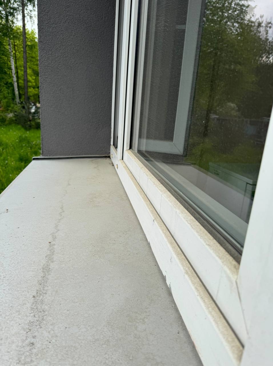 Brudny parapet i rama okienna z widocznymi zaciekami i zabrudzeniami. Widok z boku, zieleń w tle.