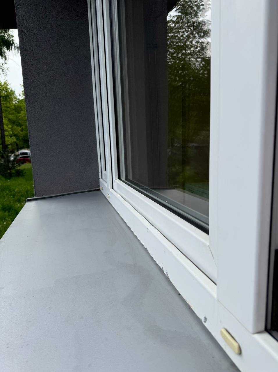 Białe okno PCV z widocznymi zabrudzeniami i odpryskami farby na ramie oraz brudnym parapetem zewnętrznym. Widoczne odbicie zieleni w szybie.