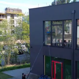 Pracownik myje okna na wysokości przy użyciu specjalistycznego sprzętu teleskopowego na tle szarej elewacji budynku i zieleni. Widoczny fragment budynku w tle.