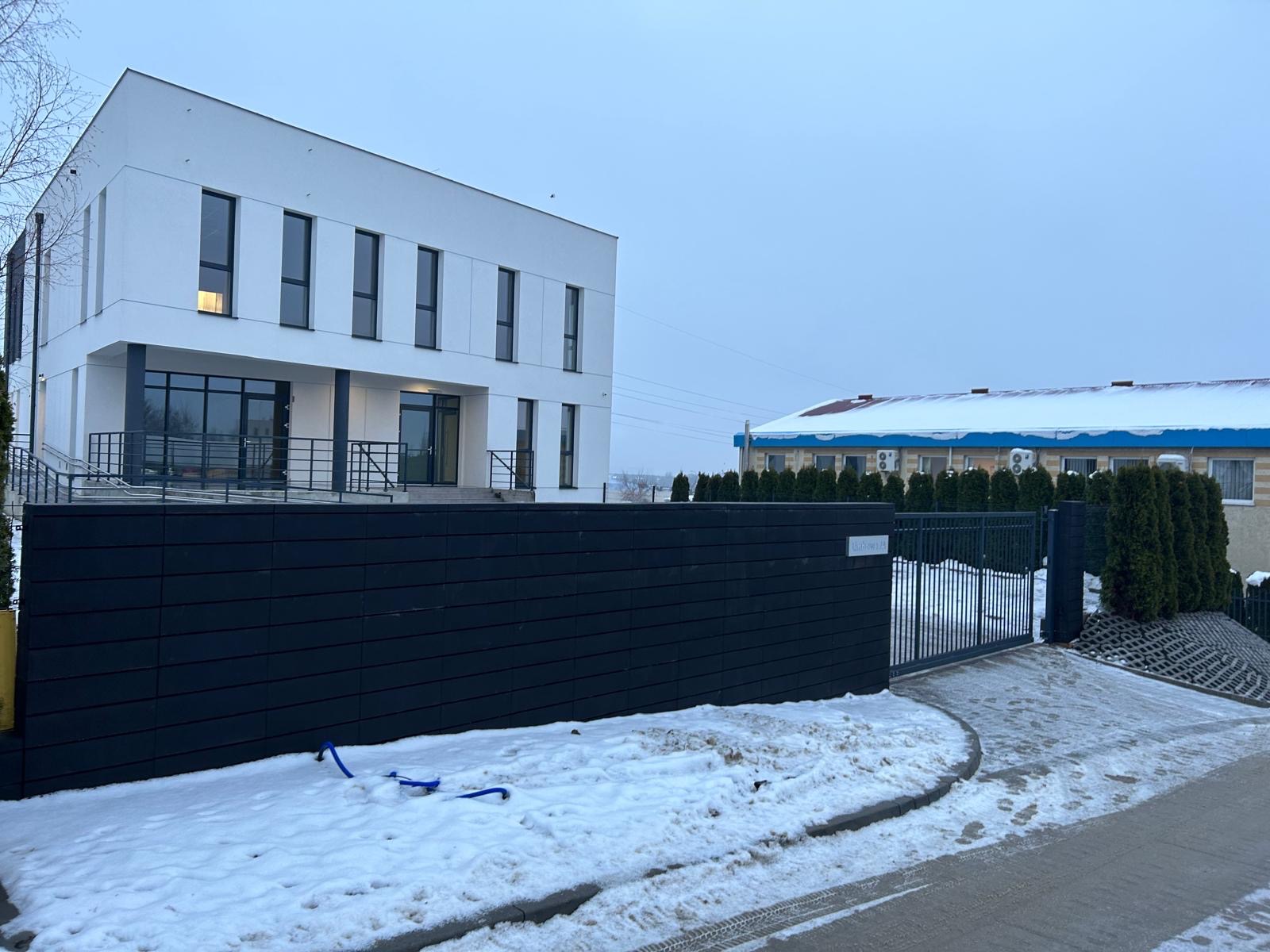 Biały, dwukondygnacyjny budynek z czarnymi ramami okiennymi i balustradami, widoczny zza czarnego ogrodzenia w zimowej scenerii. W tle budynek z niebieskim dachem.