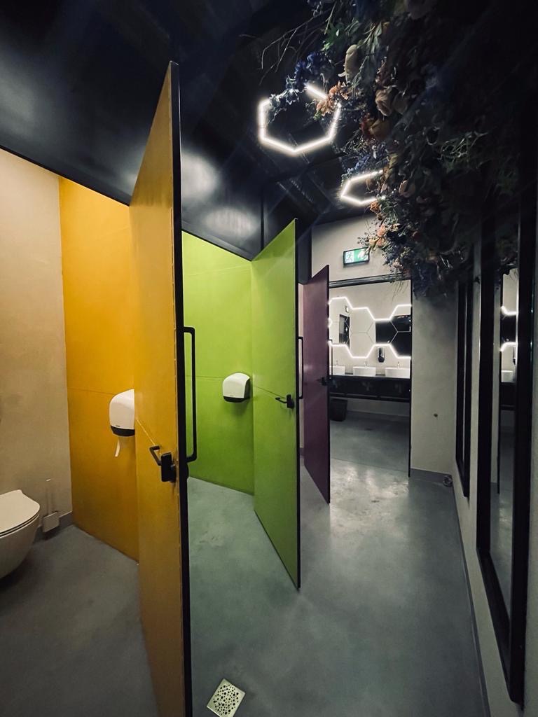 Kolorowe kabiny toaletowe z otwartymi drzwiami (żółte, zielone, fioletowe), dekoracje roślinne pod sufitem i sześciokątne lampy w nowoczesnym wnętrzu.