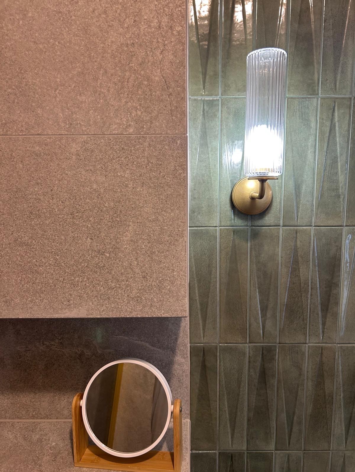 Wykończona ściana łazienki z beżowych i zielonych płytek. Na zielonej ścianie lampa ścienna, a na beżowej małe okrągłe lusterko na drewnianej podstawie.