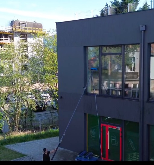 Pracownik myje okna na wysokości przy użyciu specjalistycznego sprzętu teleskopowego na tle szarej elewacji budynku i zieleni. Widoczny fragment budynku w tle.