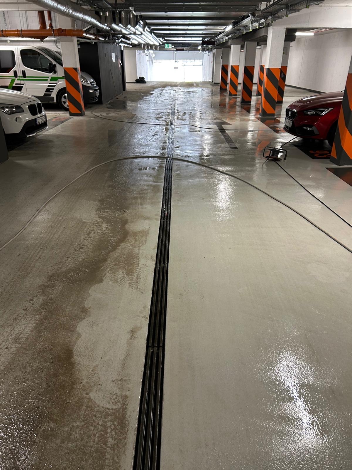 Mycie posadzki w garażu podziemnym. Widoczny efekt czyszczenia: lewa strona brudna, prawa umyta. W tle słupy z pomarańczowymi pasami i zaparkowane auta.