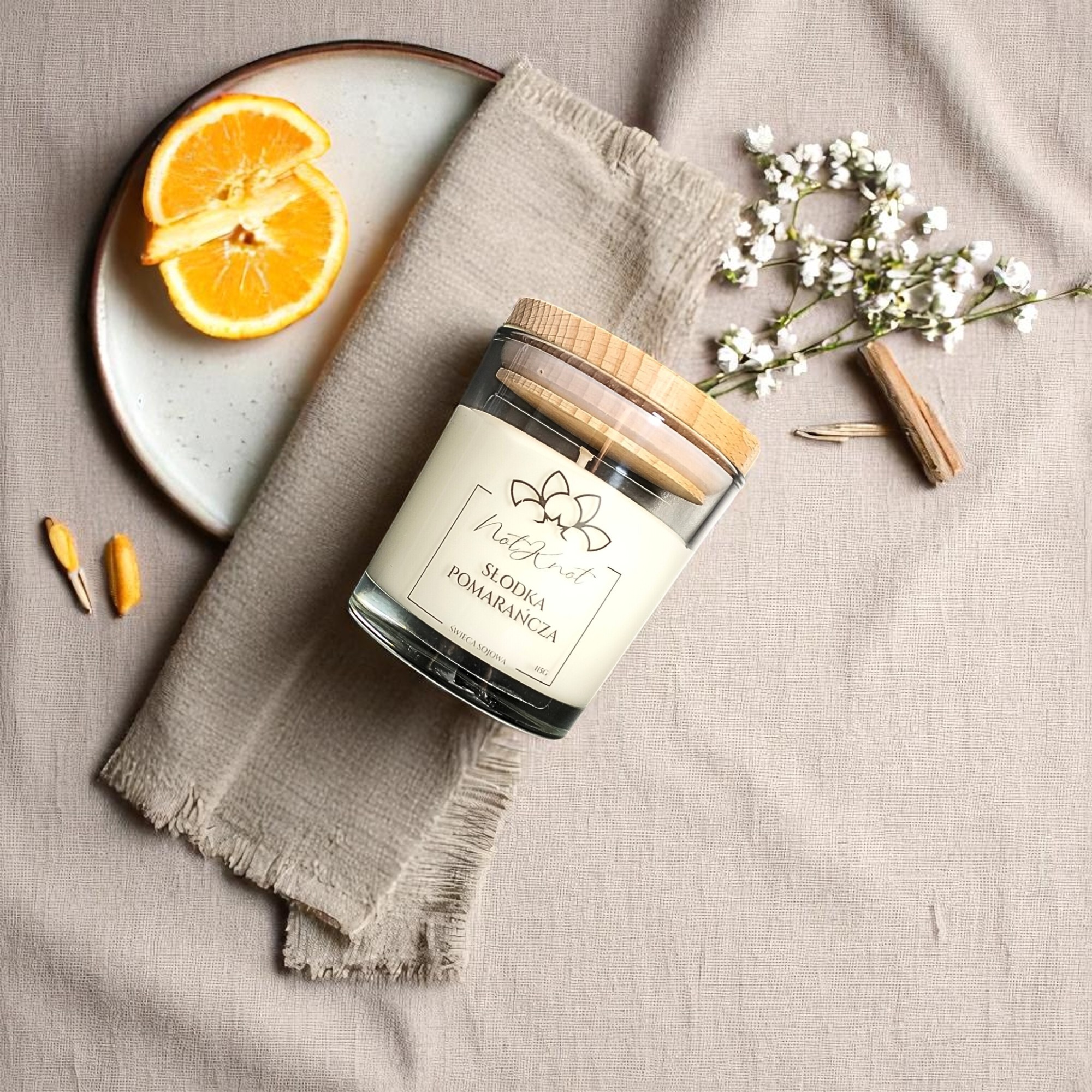 Świeca sojowa 'Słodka Pomarańcza' w szklanym słoiku z drewnianym wieczkiem, plaster pomarańczy na talerzu, gałązka kwiatów i laska cynamonu na beżowym tle.