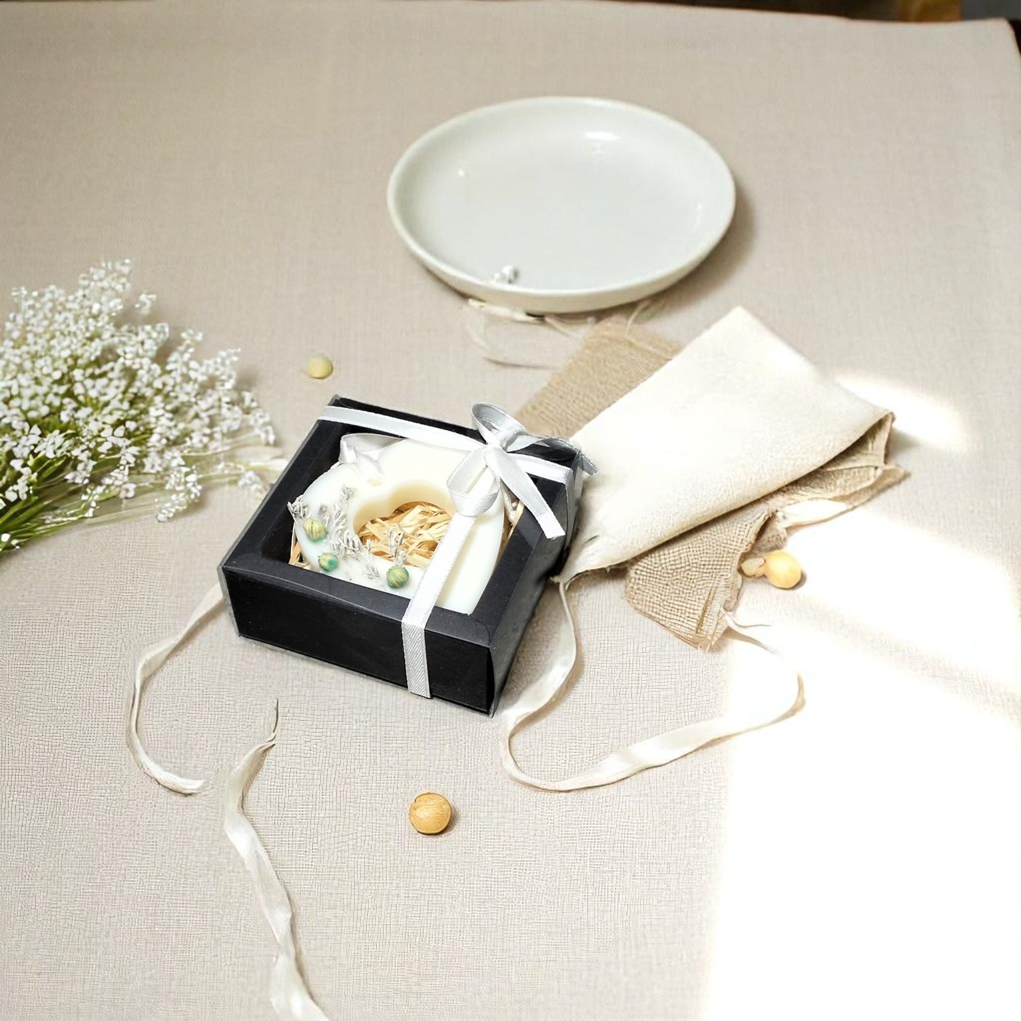 Ręcznie robione mydło w kształcie serca w czarnym pudełku z kokardą, obok woreczek jutowy i gipsówka na beżowym tle.