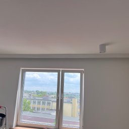 Usługi budowlane illia Zakhariv - Białe wnętrze po malowaniu ścian z oknem i widokiem na miasto. Drabina malarska z wiadrem w rogu. Nowoczesne oświetlenie sufitowe punktowe.