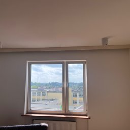 Usługi budowlane illia Zakhariv - Jasne wnętrze z oknem na budynek szkolny. Dwa okrągłe, białe reflektory sufitowe. Świeżo pomalowane ściany w odcieniach bieli i beżu. Grzejnik pod oknem.