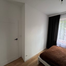 Usługi budowlane illia Zakhariv - Pokój z białymi ścianami, drewnianą podłogą i łóżkiem z brązową narzutą przy oknie z czarnymi i białymi zasłonami. Widoczna biała, zamknięta  płycinowa drzwi z czarną klamką.