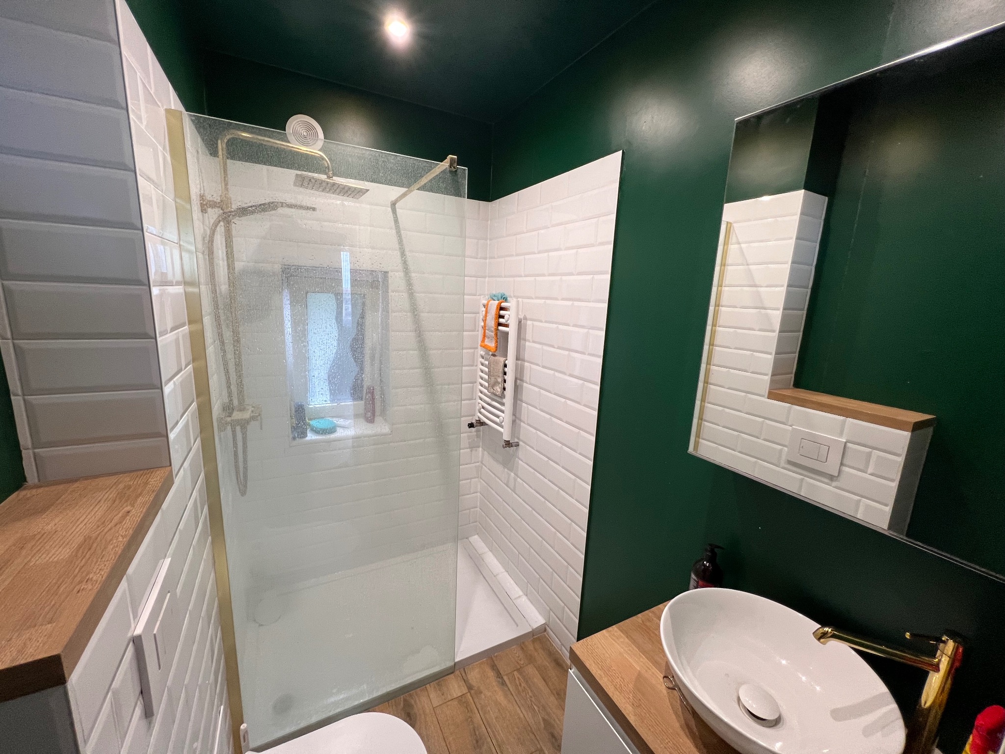 Nowoczesna łazienka z kabiną prysznicową typu walk-in, białą armaturą i drewnianymi akcentami na tle ścian w kolorze butelkowej zieleni.