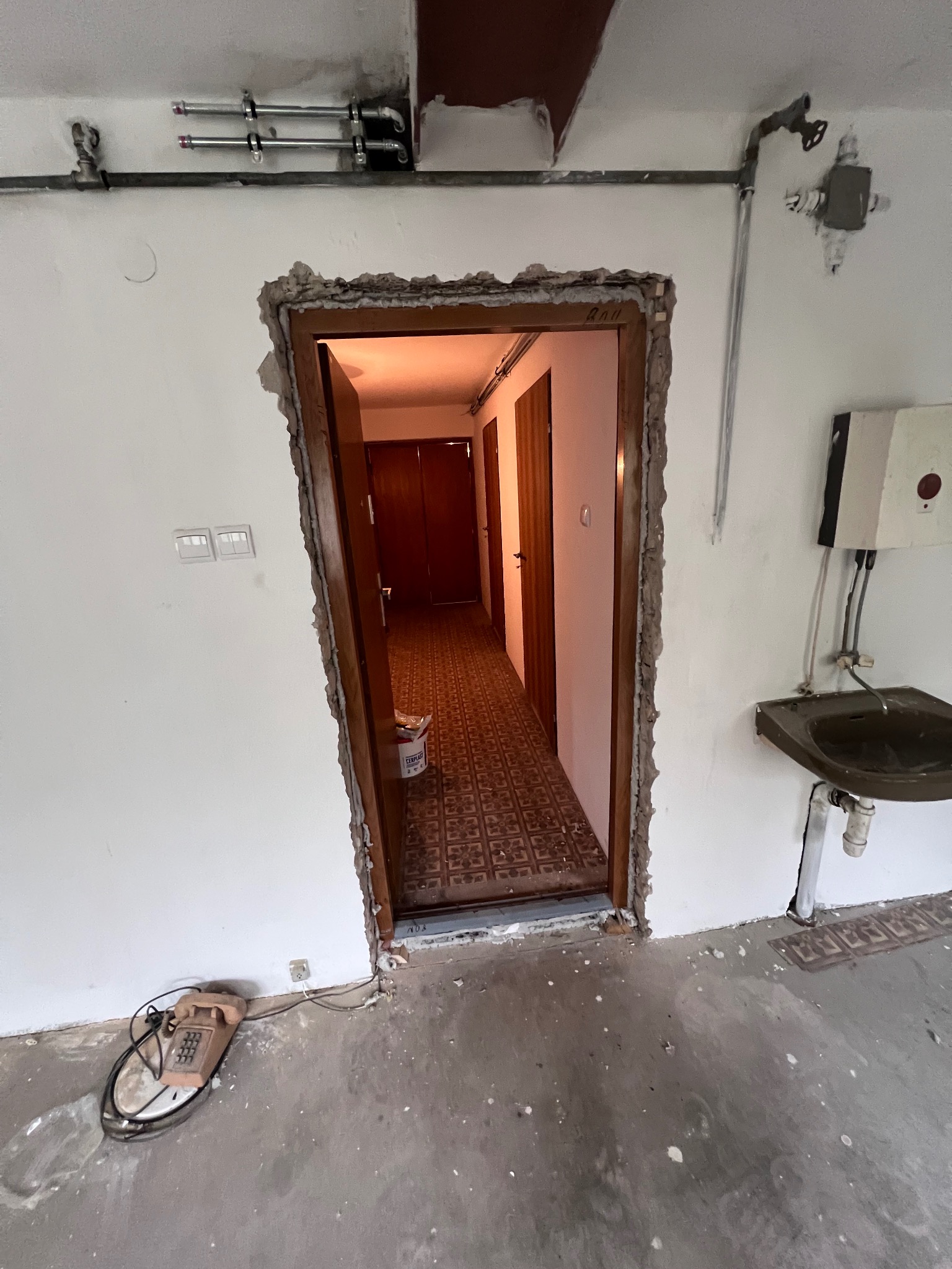 Niedokończony otwór drzwiowy w trakcie remontu. Widoczne instalacje, stara umywalka i telefon stacjonarny. Podłoga w trakcie przygotowania.